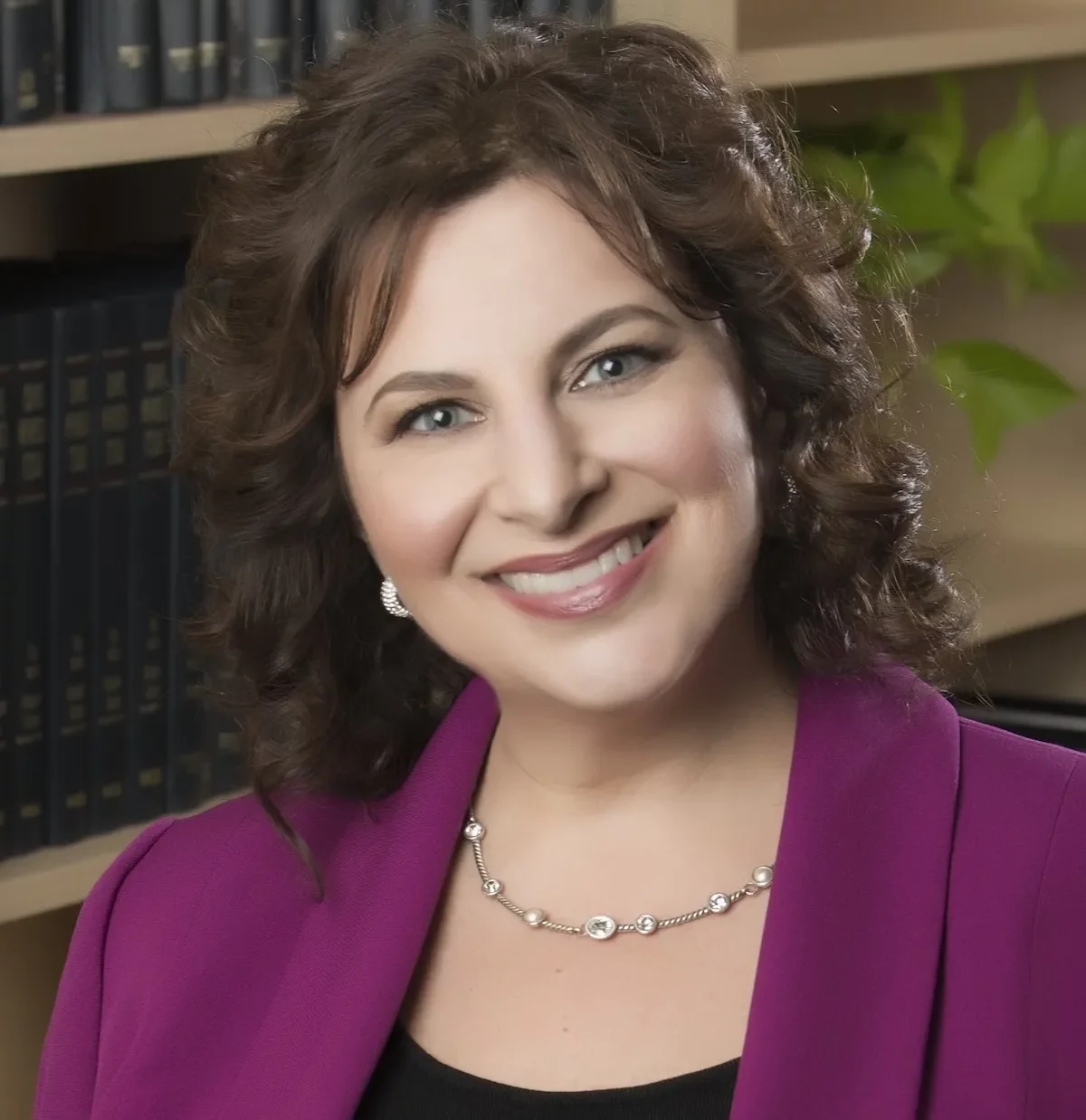 roni-sokol-pink-jacket-croppedv3-1200w Roni Sokol smiling at the camera in front of cubed shelves full of law books. She is wearing a black scoop necked dress under a fuscia jacket, and a thin gold and white necklace. She has green eyes and brown wavy shoulder length hair.